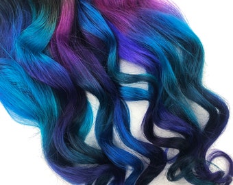 Può includere: Un'estensione per capelli lunga e ondulata con un gradiente di colori, che parte dal nero alle radici, passando al viola, poi al blu e infine al blu-verde alle punte.