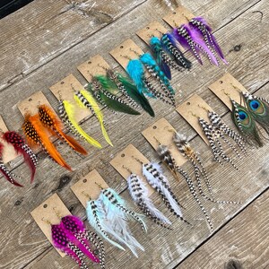May include: Assortment of feather earrings displayed on brown wooden boards. The earrings feature various colors and patterns, including peacock, red, orange, yellow, green, blue, and white.