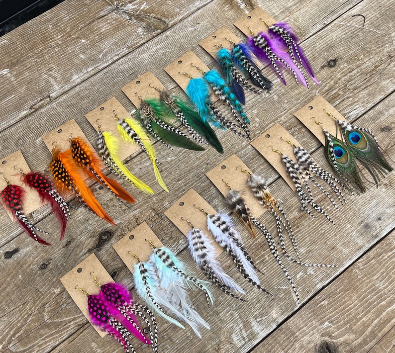 May include: Assortment of feather earrings displayed on brown wooden boards. The earrings feature various colors and patterns, including peacock, red, orange, yellow, green, blue, and white.