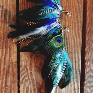 Puede incluir: Un brazalete de oreja artesanal con plumas azules, turquesas, negras y blancas. El dise&ntilde;o incorpora plumas de pavo real y detalles de alambre, creando un accesorio &uacute;nico. Sobre fondo de madera.