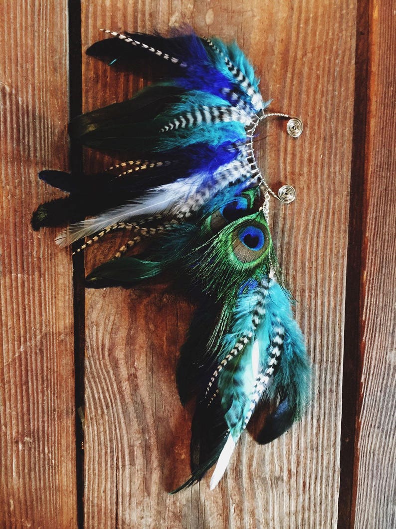 Puede incluir: Un brazalete de oreja artesanal con plumas azules, turquesas, negras y blancas. El dise&ntilde;o incorpora plumas de pavo real y detalles de alambre, creando un accesorio &uacute;nico. Sobre fondo de madera.