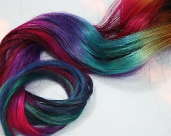 May include: Close-up of colorful hair extensions in shades of red, pink, purple, teal, and gold. The hair is styled in a wavy pattern, with the colors blending together. The background is plain white.