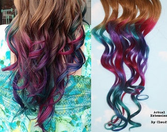 May include: A set of clip-in hair extensions with a gradient of colors, starting with a light brown at the top and transitioning to teal, purple, and pink at the bottom. The extensions are wavy and have a natural look.