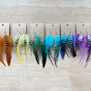 May include: A set of 10 pairs of feather earrings. Each pair features a different color combination of feathers, including red, orange, yellow, green, blue, purple, and pink. The feathers are arranged in a layered design, with the longest feathers at the bottom and the shortest feathers at the top. The earrings are attached to gold-colored hooks.