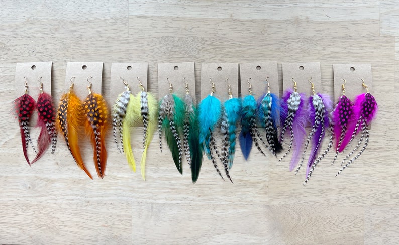 May include: A set of 10 pairs of feather earrings. Each pair features a different color combination of feathers, including red, orange, yellow, green, blue, purple, and pink. The feathers are arranged in a layered design, with the longest feathers at the bottom and the shortest feathers at the top. The earrings are attached to gold-colored hooks.