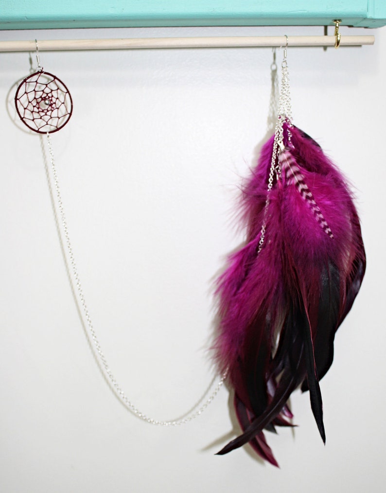 May include: A pair of earrings with a silver chain and a pink and black feather design. The earrings are hanging from a wooden rod.