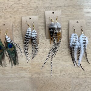 May include: Four pairs of feather earrings displayed on brown card stock. The earrings are made with various feathers, including peacock, black and white, and brown and white. The earrings are all about 4 inches long.