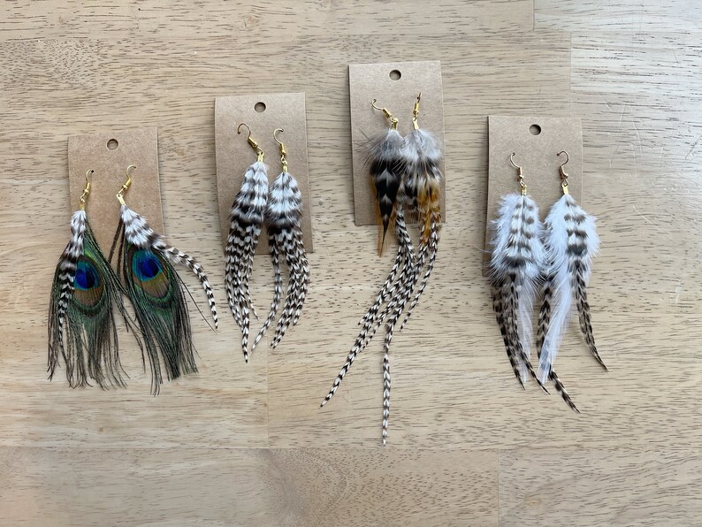 May include: Four pairs of feather earrings displayed on brown card stock. The earrings are made with various feathers, including peacock, black and white, and brown and white. The earrings are all about 4 inches long.