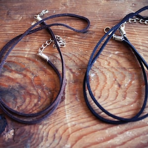 Könnte beinhalten: Zwei Choker-Halsketten aus Wildleder mit silberfarbenen Kettenverlängerungen und Verschlüssen. Eine Halskette ist dunkelbraun, die andere ist schwarz. Beide liegen auf einer Holzoberfläche mit sichtbarer Maserung.