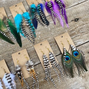 May include: Assortment of feather earrings displayed on brown card stock. The earrings feature various colors and patterns, including teal, purple, black and white, and peacock feathers.