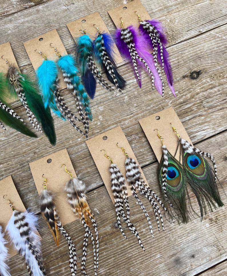 May include: Assortment of feather earrings displayed on brown card stock. The earrings feature various colors and patterns, including teal, purple, black and white, and peacock feathers.
