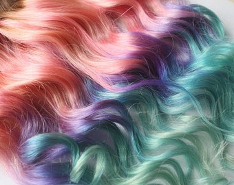 May include: Close-up of wavy, colorful hair extensions. The hair transitions from shades of peach and pink to purple and teal. The strands are soft and flowing, creating a vibrant and textured appearance. Ideal for cosplay or adding a pop of color.