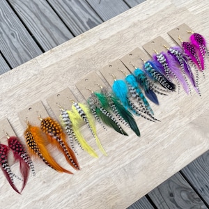 May include: A set of 10 pairs of feather earrings in various colors, including red, orange, yellow, green, blue, purple, and pink. The earrings are displayed on a wooden surface.