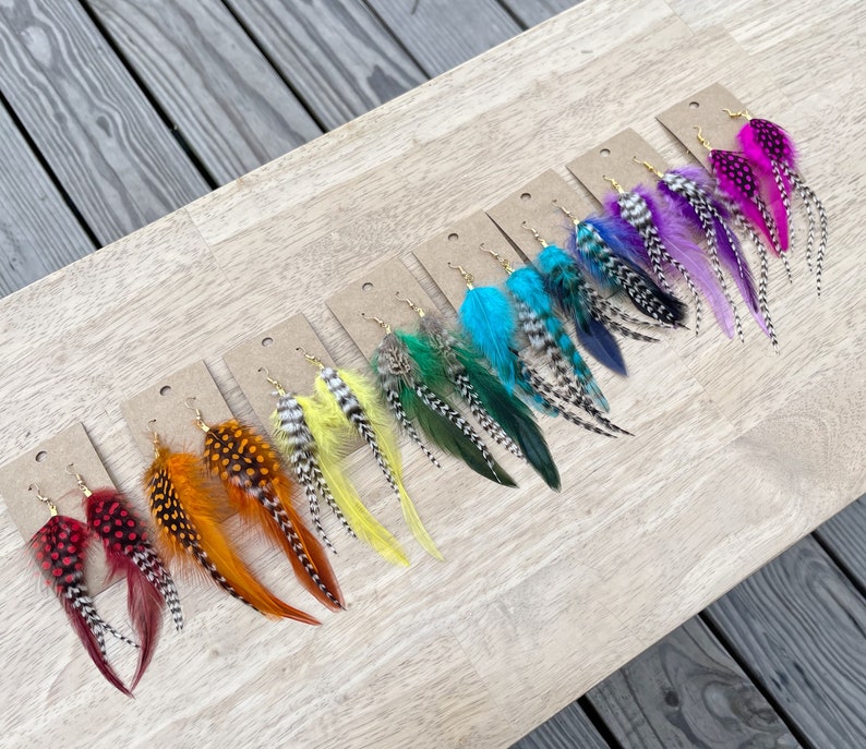 May include: A set of 10 pairs of feather earrings in various colors, including red, orange, yellow, green, blue, purple, and pink. The earrings are displayed on a wooden surface.