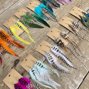 May include: Assortment of feather earrings displayed on brown card stock. The earrings feature various colors and patterns, including turquoise, green, yellow, orange, pink, white, and peacock feathers.