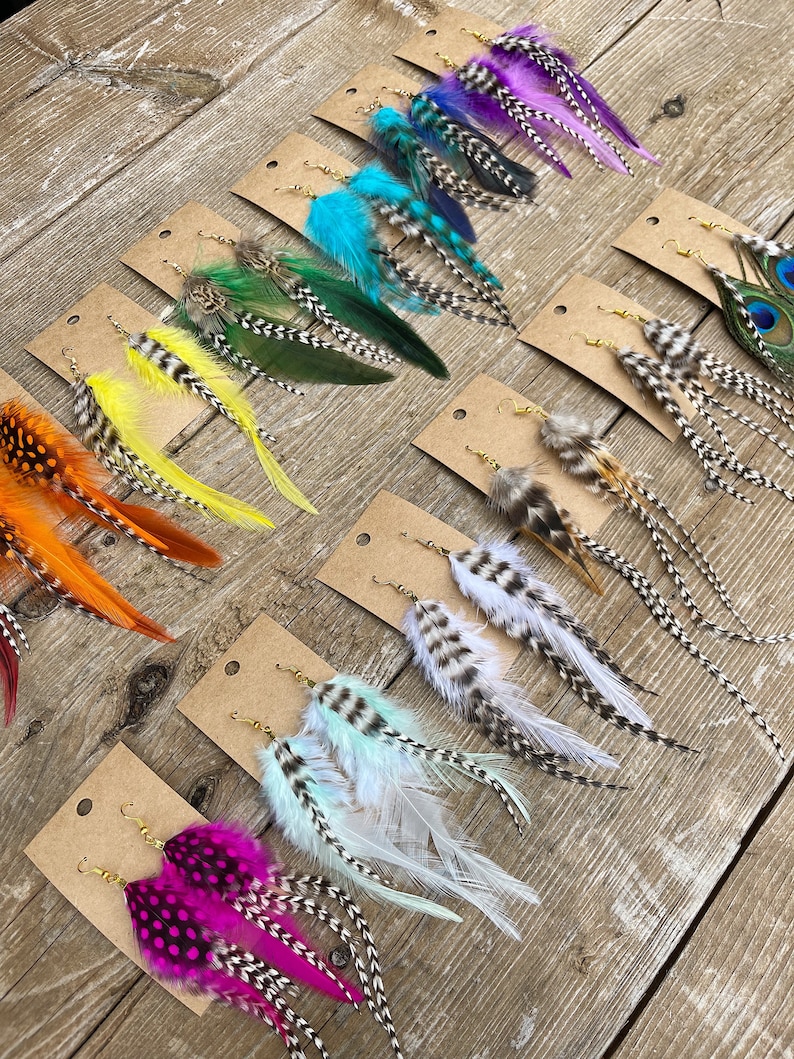 May include: Assortment of feather earrings displayed on brown card stock. The earrings feature various colors and patterns, including turquoise, green, yellow, orange, pink, white, and peacock feathers.
