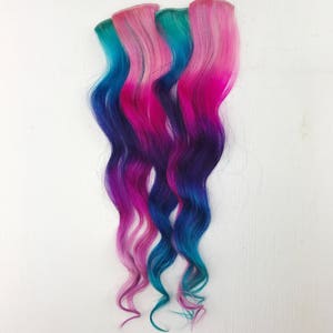 May include: Three strands of wavy hair extensions in shades of pink, blue and purple. The hair is styled in a loose wave.