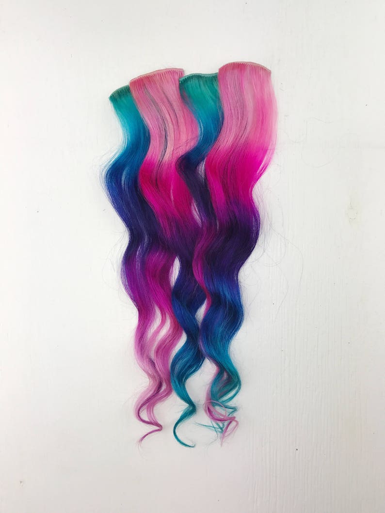 May include: Three strands of wavy hair extensions in shades of pink, blue and purple. The hair is styled in a loose wave.