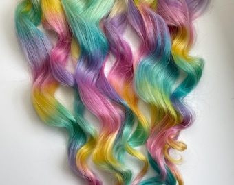 May include: A set of wavy blonde hair extensions with a rainbow ombre color effect. The hair is dyed in shades of yellow, green, blue, purple, and pink.