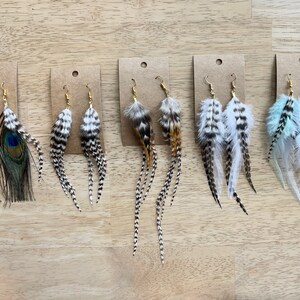 May include: Six pairs of feather earrings displayed on brown card stock. The earrings are made with various feathers, including peacock, white, and black feathers. The earrings are all different lengths and styles.