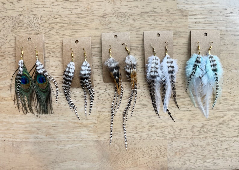 May include: Six pairs of feather earrings displayed on brown card stock. The earrings are made with various feathers, including peacock, white, and black feathers. The earrings are all different lengths and styles.