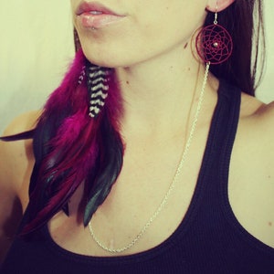 May include: A woman wearing a black tank top with a silver chain necklace and a pair of long, dangling earrings. The earrings are made of red thread and feature a circular design. The woman is also wearing a feather earring with pink, black, and white feathers.