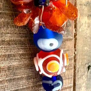 Könnte beinhalten: Dekorativer Hängeornament mit bernsteinfarbenen, unregelmäßig geformten Steinen und bunten Glasperlen. Die Perlen umfassen ein blaues Auge, ein rot-weißes Design und eine blaue Perle mit weißen Wirbeln. Das Ornament ist auf Draht aufgefädelt.