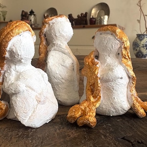 Puede incluir: Tres esculturas hechas a mano de figuras con cuerpos blancos y cabello dorado. Las esculturas están dispuestas sobre una superficie de madera. Las figuras tienen una apariencia texturizada y están sentadas.