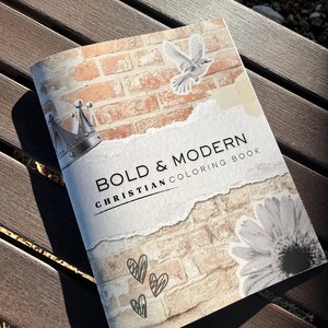 May include: A "Bold & Modern Christian Coloring Book" lies on a wooden surface. The cover features a brick wall design, a dove, a crown, a daisy, and three sketched hearts. The title is printed in black, with the words "Bold & Modern" in a large font.