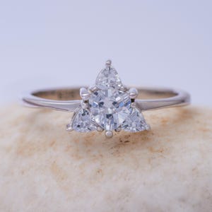 May include: A silver ring featuring a central triangular-cut diamond, flanked by two smaller triangular diamonds. The ring is displayed on a light-toned surface, highlighting the brilliance of the gemstones. The band is slender and polished.
