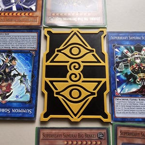 Puede incluir: Una colección de cartas coleccionables de Yu-Gi-Oh! y un tarjetero negro y dorado. Las cartas presentan varios personajes y texto, incluyendo "Summon Sorceress" y "Superheavy Samurai Scarecrow". El tarjetero tiene un diseño de ojo.