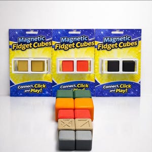 Può includere: Tre confezioni di Magnetic Fidget Cubes con cubi dorati, rossi e neri. La confezione blu presenta dettagli gialli e il testo "Connect, Click and Play!". Cubi aggiuntivi in verde, arancione, beige e grigio sono impilati sotto.