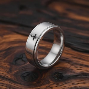 Peut inclure: Une bague argentée ornée d'une croix noire. La bague présente une finition polie et un design épuré. La bague est présentée sur une surface en bois.