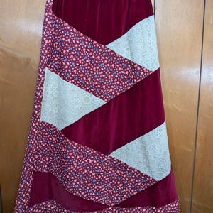 May include: A floor-length skirt featuring a patchwork design of deep red velvet, cream-colored lace panels, and a dark fabric with a small floral print in shades of pink, blue, and white. The skirt has an elastic waistband and a flared silhouette.
