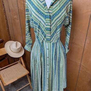 May include: A long-sleeved button-down dress featuring a vibrant pattern of vertical and diagonal stripes in shades of green, blue, and cream. The garment has a collared neckline, a full-length front button placket, and a pleated skirt.