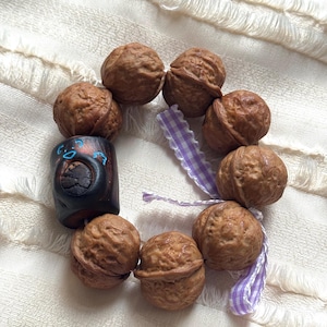 May include: A handmade bracelet featuring brown walnuts and a dark wooden bead with blue and black accents. The bracelet is accented with a purple and white checkered ribbon. The bracelet is displayed on a white, textured surface.
