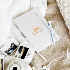May include: A white journal with the text "be the light" and a sunrise graphic. A light blue pen rests on the journal, and several small photographs and a camera are nearby. The scene is set on a soft, textured surface.