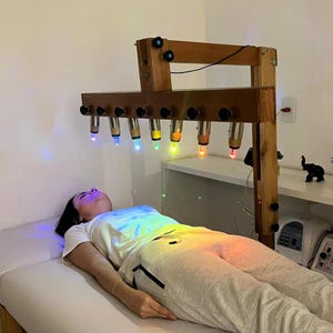 以下が含まれることがあります： 青、黄、オレンジ、赤の光を放つ7つの電球を備えた木製の光療法装置。この装置は、黒いノブが付いた頑丈な木製フレームに取り付けられ、白い施術用ベッドの上に配置されています。