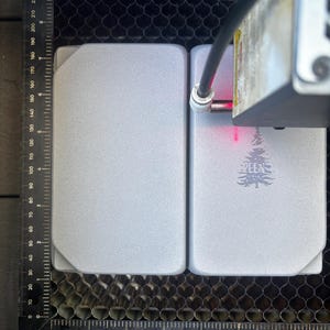 Może przedstawiać: Dwa srebrne prostokątne obiekty z zaokrąglonymi rogami umieszczone są na powierzchni o strukturze plastra miodu. Używany jest grawer laserowy, widoczna jest czerwona wiązka lasera. Jeden obiekt ma wzór drzewa i wygrawerowane słowo "REEN". Z boku znajduje się linijka.