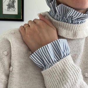 Puede incluir: Suéter color crema con cuello alto y puños acanalados. El suéter presenta un cuello y puños con volantes a rayas azules y blancas. Se ve un anillo de oro en la mano que descansa sobre el suéter.