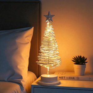 Könnte beinhalten: Dekorative Weihnachtsbaum-Lampe mit silbernem Drahtrahmen und warmweißen LED-Lichtern. Der Baum wird von einem silbernen Stern gekrönt und steht auf einem weißen Sockel. Eine kleine Topfpflanze und ein Buch befinden sich neben der Lampe.