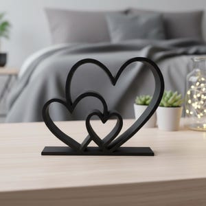 May include: A black heart-shaped sculpture featuring three interconnected hearts of varying sizes. The sculpture is made of a solid material and stands on a rectangular base. The decor is placed on a light-colored wooden surface.
