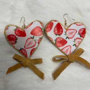 May include: A pair of heart-shaped earrings with a strawberry print on a white backdrop. The edges are stitched with gold thread, and each earring has a small, tan bow. The earrings hang from gold-coloured hooks.