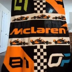 Pode incluir: Um grande cobertor de malha com o logotipo da McLaren e imagens de carros de corrida. O cobertor tem um fundo preto com detalhes em laranja, branco e verde. A palavra "McLaren" é exibida em laranja.