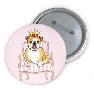 Puede incluir: Un botón redondo con un bulldog que lleva una corona dorada y está sentado en una silla a rayas rosas. El texto "UGA" está en la corona. El botón tiene un fondo rosa y un reverso de alfiler plateado.