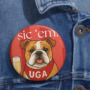 Puede incluir: Un botón redondo rojo con el texto "sic 'em." presenta una ilustración de un bulldog con un pañuelo rojo con "UGA". También se muestra una copa de champán. El botón está prendido a una chaqueta vaquera.