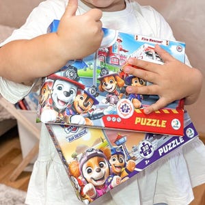 Puede incluir: Un niño sostiene tres coloridos rompecabezas de Paw Patrol de 30 piezas. Las cajas de rompecabezas presentan imágenes de perros de dibujos animados en varios escenarios de rescate, con las palabras "Fire Rescue!" y "Air Rescue" visibles. Los rompecabezas son para mayores de 3 años.