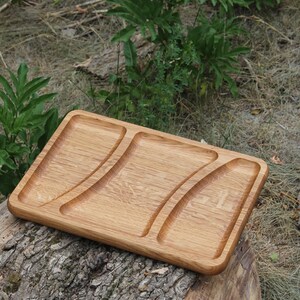 Puede incluir: Una bandeja rectangular de madera con tres compartimentos curvos. La bandeja es de madera clara y está sobre un tocón de árbol. El fondo presenta follaje verde y hierba seca.