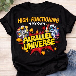 May include: Black t-shirt with the text "HIGH-FUNCTIONING IN MY OWN PARALLEL UNIVERSE" in yellow and red. The design includes two astronauts in space suits, surrounded by colorful graphics.