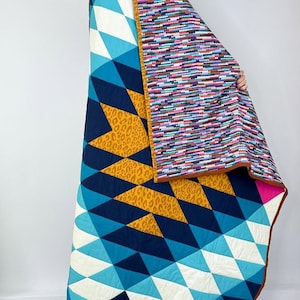 Könnte beinhalten: Ein farbenfroher Quilt mit geometrischen und Leopardenmustern. Er hat Blau-, Weiß- und Goldtöne mit einem rosa Rand. Die Rückseite hat ein Muster aus kleinen, farbigen Punkten. Der Quilt ist gefaltet und zeigt seine verschiedenen Muster.